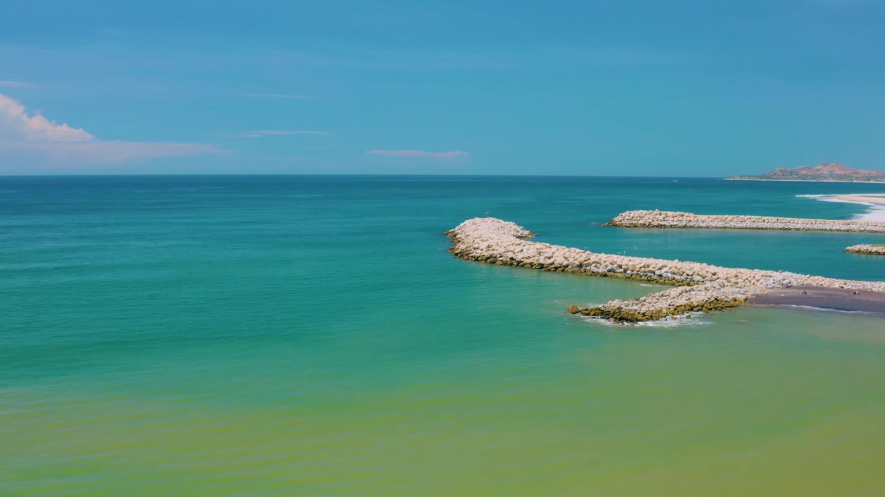 San Jose Del Cabo coastal frame with the ocean horizon running across the upper third, pale rock jetties in the midground, distant sandy coast to the right, and no visible people.