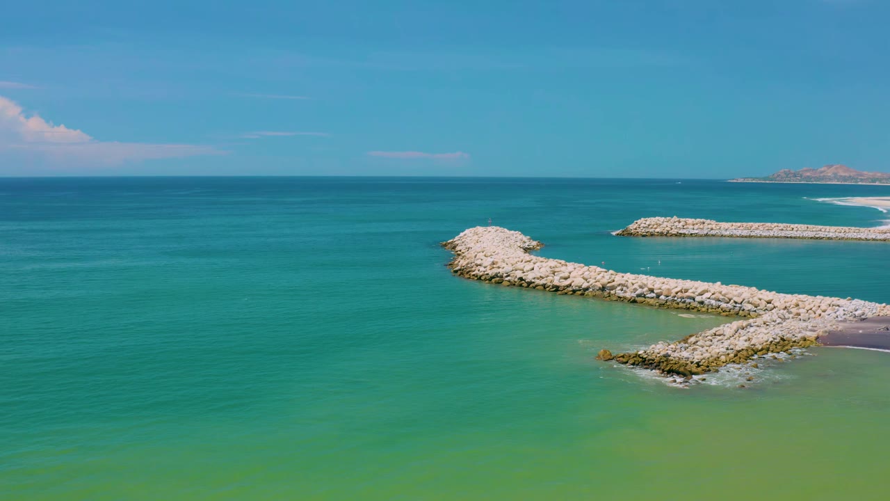 Aerial view in San Jose Del Cabo, Mexico with the curved rock arm centered lower in frame, a straighter rock barrier beyond it, and no visible people.
