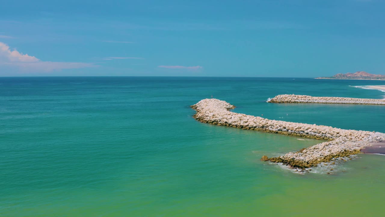 San Jose Del Cabo aerial frame with broad teal water, pale rocks forming a bay entrance, a distant sandy beach at right, and no visible people.