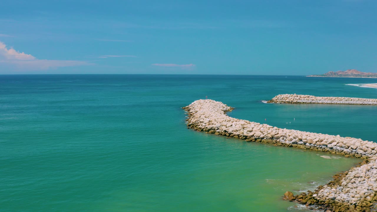 Opening aerial view of San Jose Del Cabo, Mexico with turquoise water, a curved pale rock breakwater in the foreground, a straighter breakwater beyond it, distant shoreline, and no visible people.