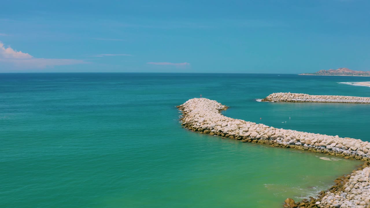 San Jose Del Cabo, Mexico coastline frame with the curved rock breakwater running across clear green-blue water, a straight offshore barrier behind it, pale shoreline at right, and no visible people.