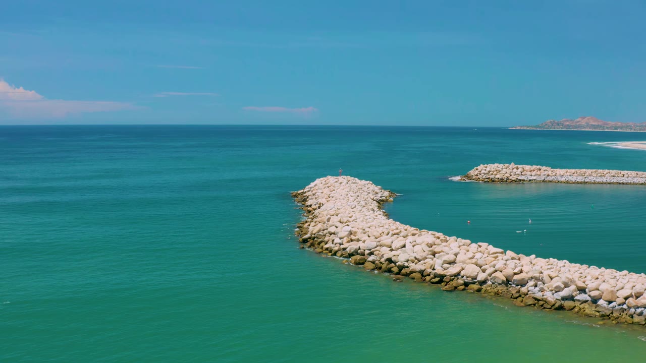 Aerial San Jose del Cabo frame with the pale curved rocks centered more strongly, calm green-blue water below, a second rock barrier at right, and no visible people.