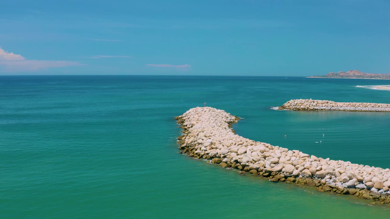 High coastal view in San Jose del Cabo, Mexico, showing the curved rock formation reaching from the lower right toward open water, distant land at the horizon, and no visible people.