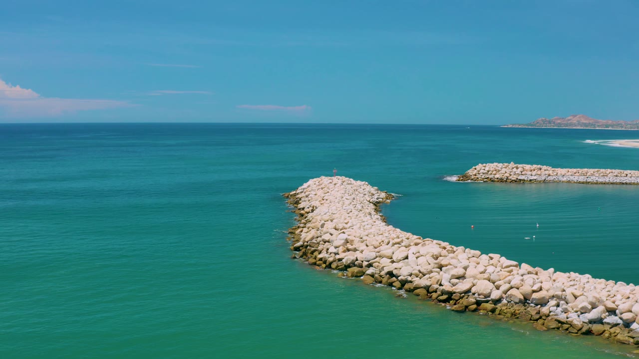 San Jose del Cabo aerial view with the curved rock formation lower in frame, calm ocean filling most of the image, a distant rocky barrier to the right, and no visible people.