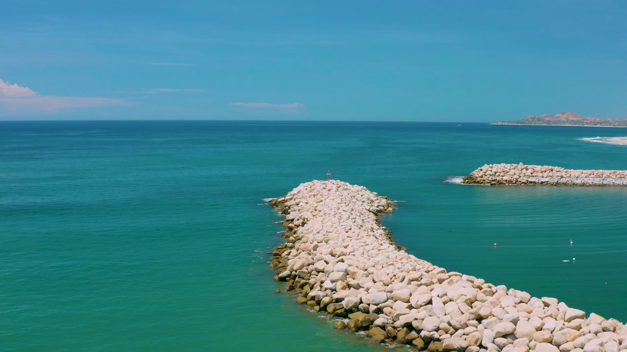 Opening frame of a pale rocky jetty extending into teal water in San Jose del Cabo, Mexico, with a distant breakwater, horizon line, and no visible people.