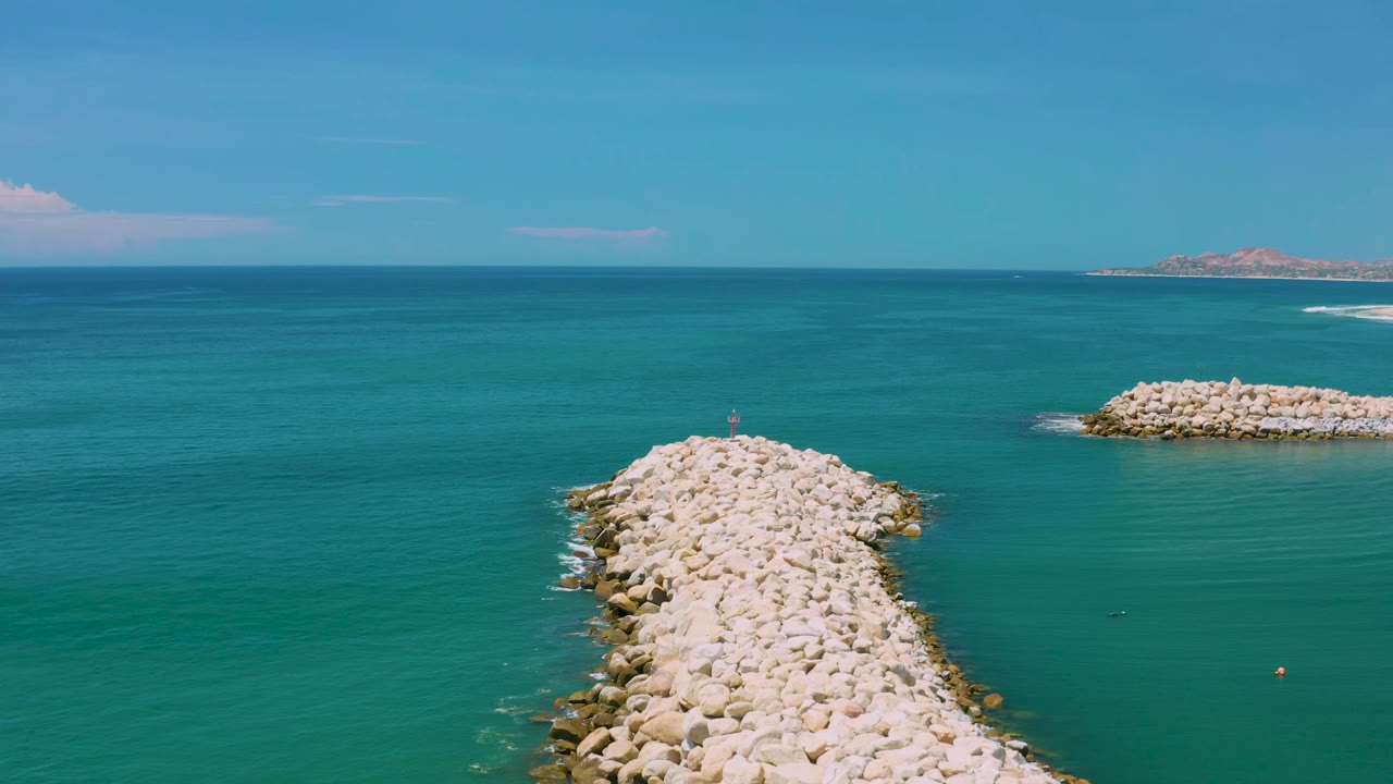 San Jose Del Cabo aerial frame showing the rock jetty lower center, blue-green water across the frame, distant land near the horizon, and no visible people.