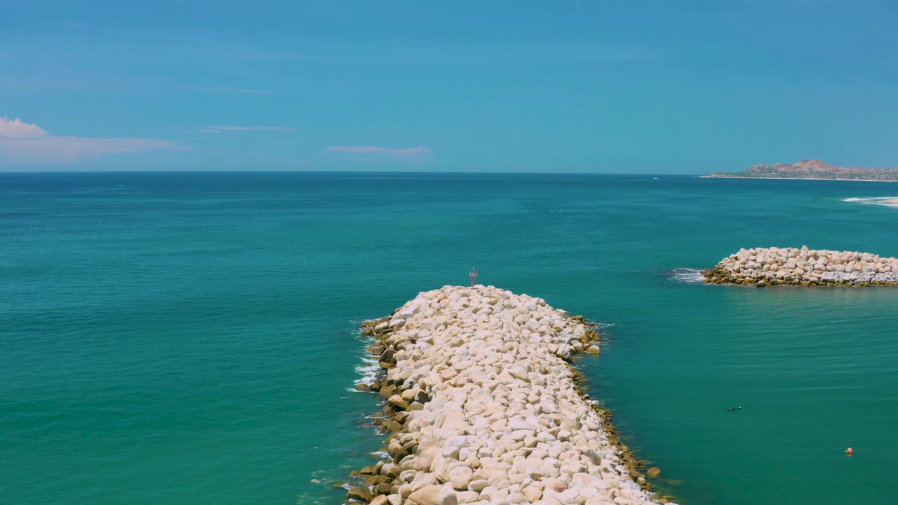 Aerial view over San Jose Del Cabo water with the pale breakwater slightly lower in frame, a second rocky structure to the right, distant land, and no visible people.