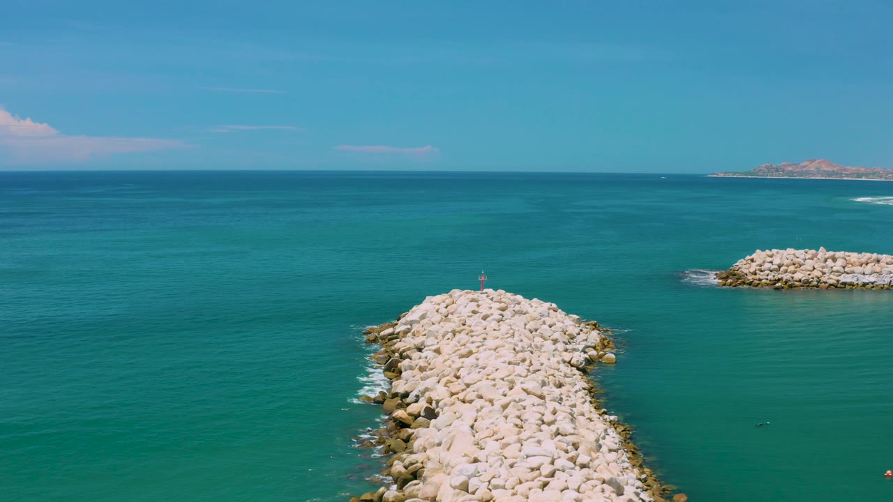 San Jose Del Cabo aerial frame with the rock breakwater rising from the bottom edge, small white wave lines along the rocks, distant land, and no visible people.