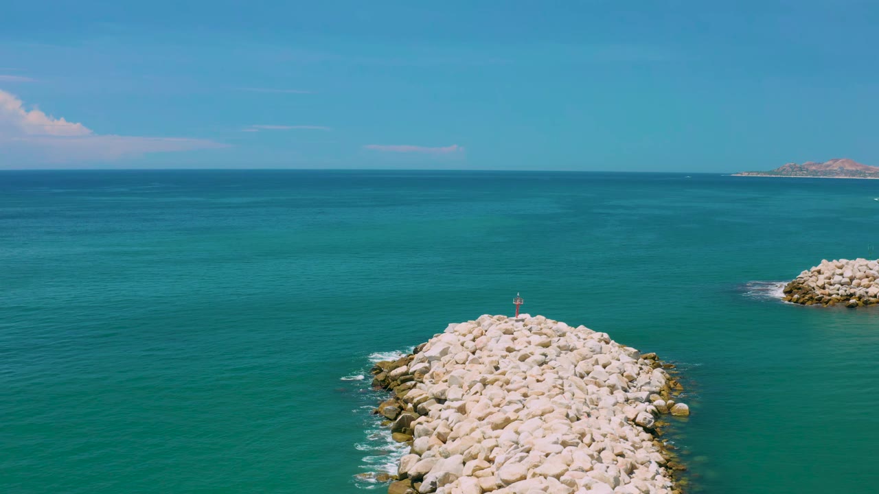 San Jose Del Cabo coastal frame with the rock breakwater centered low in view, small waves touching both sides, distant land on the right horizon, and no visible people.