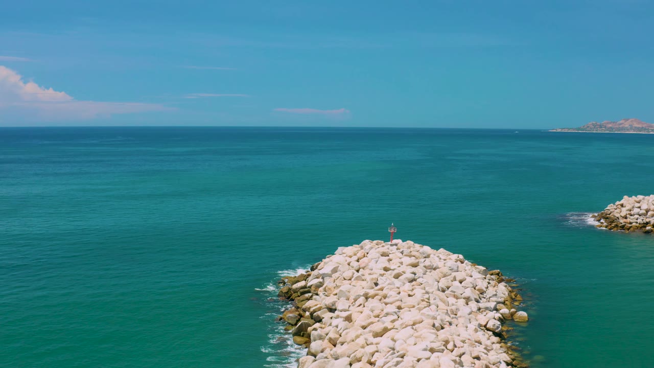 San Jose Del Cabo ocean scene with the rock breakwater slightly lower in frame, a small upright marker near its end, clear blue-green water, and no visible people.