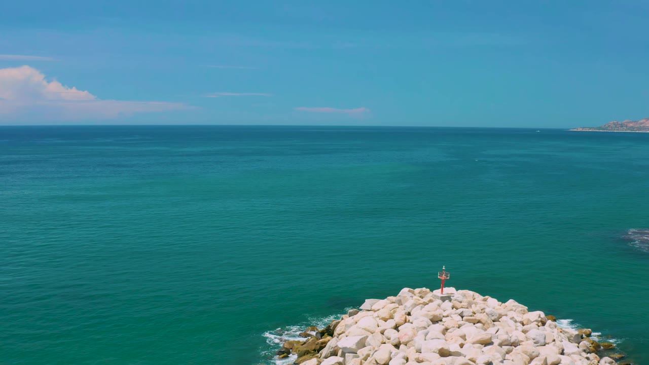 Aerial frame in San Jose Del Cabo, Mexico, showing turquoise water, the rocky breakwater lower in frame, a small red marker near center bottom, faint distant coastline, and no visible people.