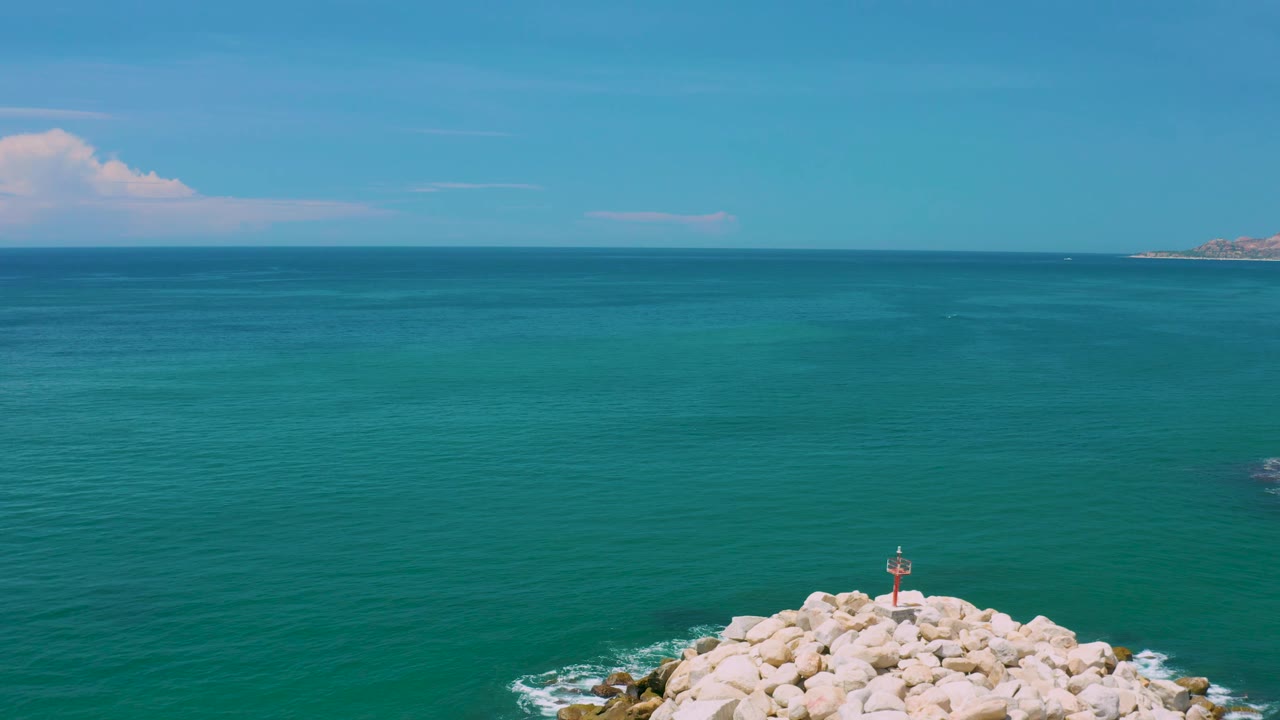 Wide aerial view off San Jose Del Cabo, Mexico, with blue-green sea across the frame, white rocks at the bottom edge, a small navigation light on the breakwater, distant land at right, and no visible people.