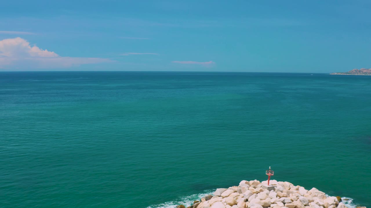 San Jose Del Cabo aerial view with calm blue water filling most of the image, a rocky jetty and red marker touching the lower edge, distant coastline on the horizon, and no visible people.