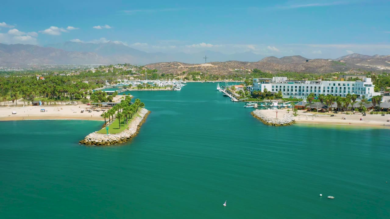 Aerial view in San Jose del Cabo, Mexico over turquoise water toward a white beachfront resort, palm-lined jetties, marina slips, low hills, and no visible people.