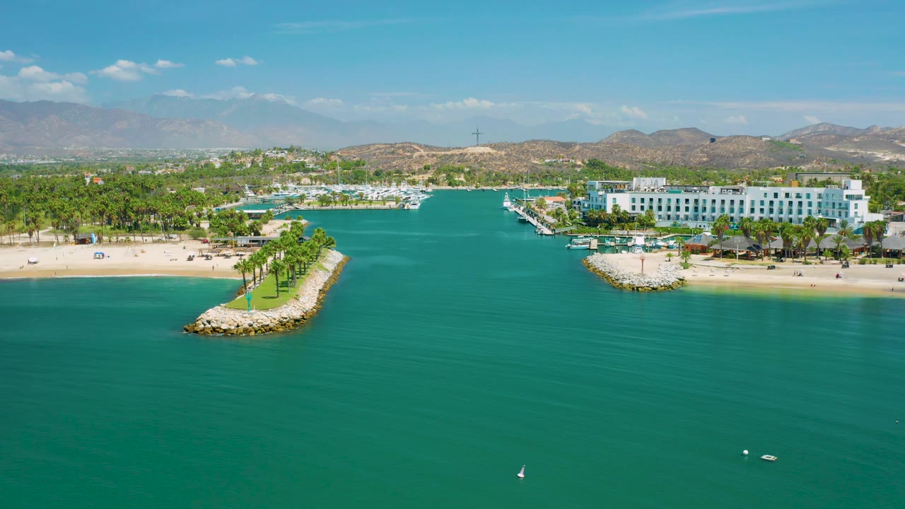 Aerial shoreline view in San Jose del Cabo, Mexico with turquoise bay water in front, pale beach, white hotel complex, marina docks, palm rows, brown hills, and no visible people.