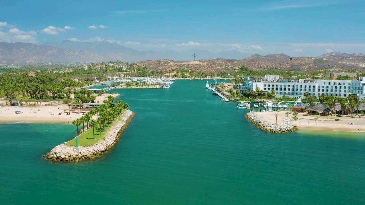 San Jose Del Cabo aerial view with the palm-lined rock jetty more centered, turquoise water in front, resort buildings along the shore, distant hills, and no visible people.