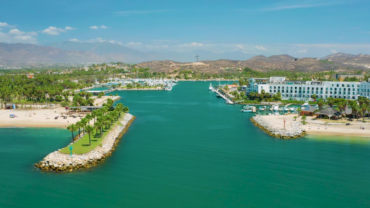 Wide San Jose Del Cabo aerial frame looking across green-blue water toward beach palms, docked boats, white resort structures, dry hills, and no visible people.