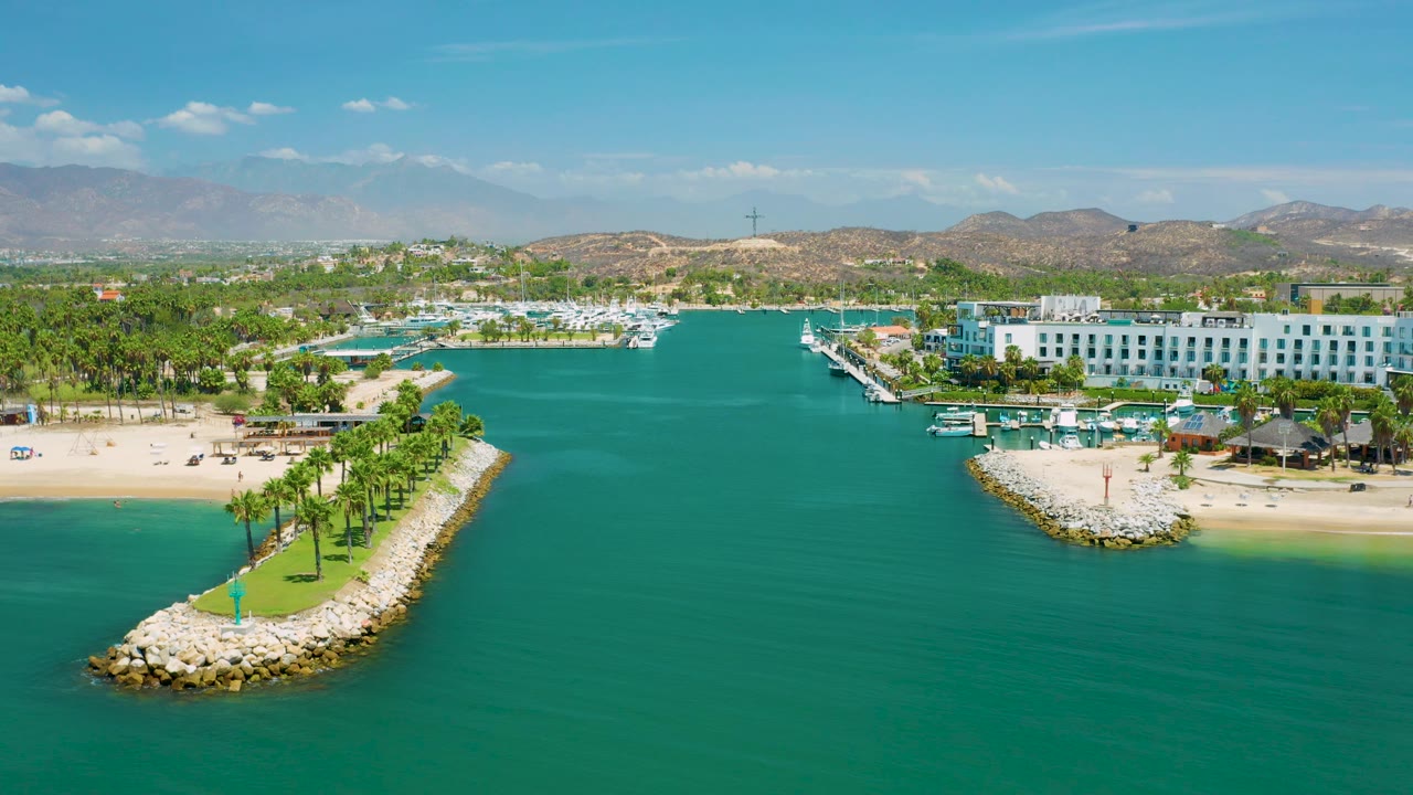 San Jose Del Cabo aerial view with the left beach and palm jetty visible, resort buildings and boats across the shore, hazy hills behind them, and no visible people.