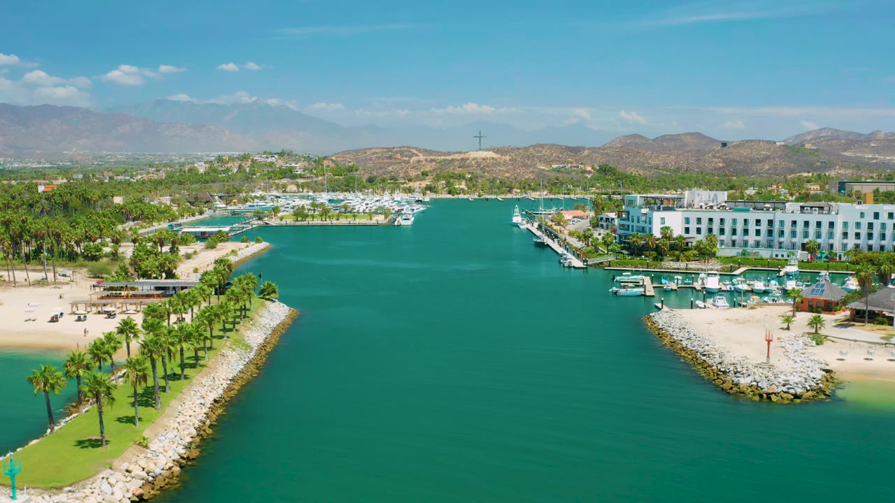 One second into the San Jose Del Cabo, Mexico clip, the aerial view holds green-blue water in the center with rock breakwaters, palms, marina slips, white resort buildings, hills, and no visible people.