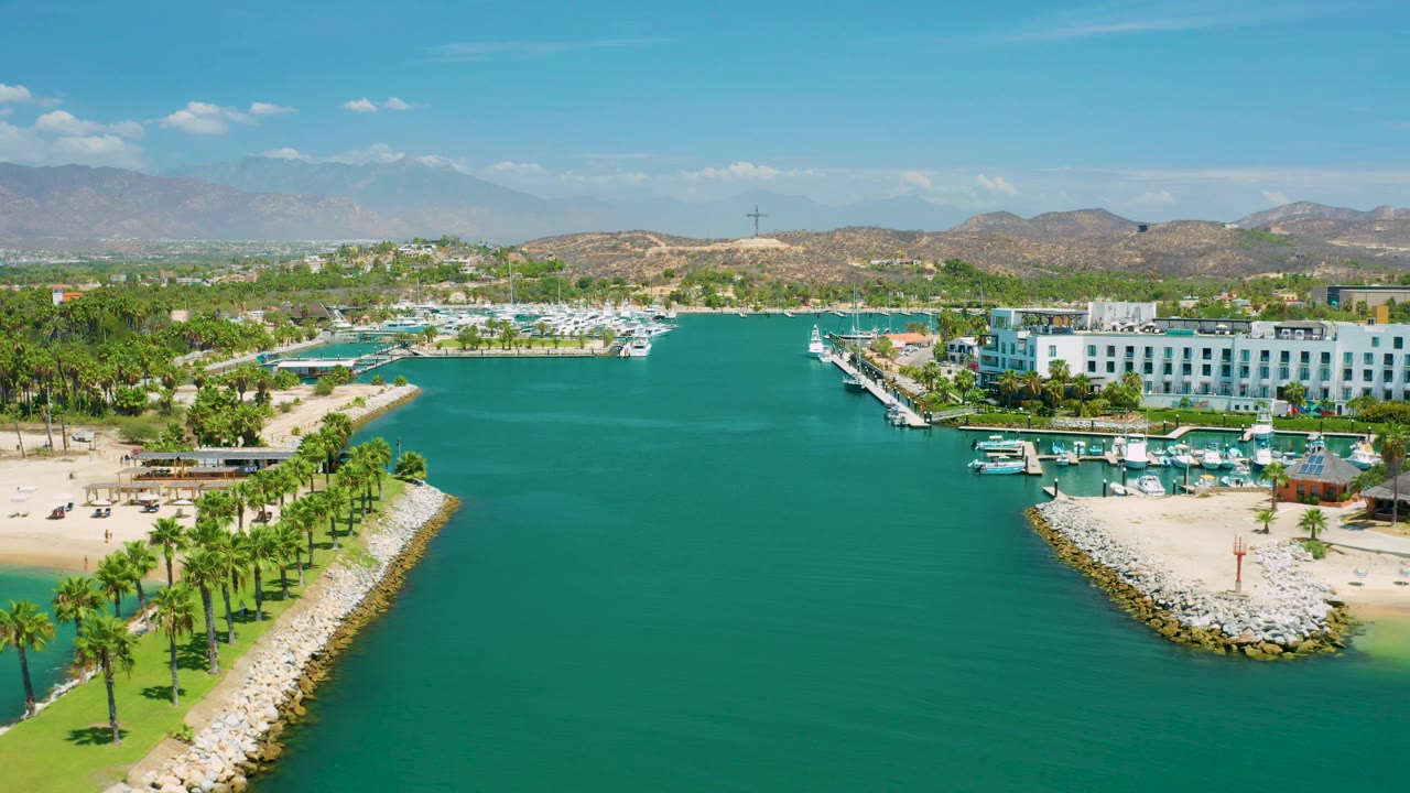 At 0:02, the San Jose Del Cabo, Mexico aerial view shows more of the right-side resort facade and marina boats beside turquoise water, sandy beach, palm rows, hills, mountains, and no visible people.