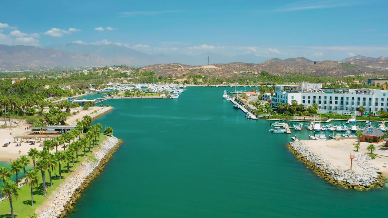 At 0:03, the wide San Jose Del Cabo, Mexico drone view keeps the inlet broad and centered, with sandy beaches on both sides, palm lines, marina boats, white buildings, distant mountains, and no visible people.