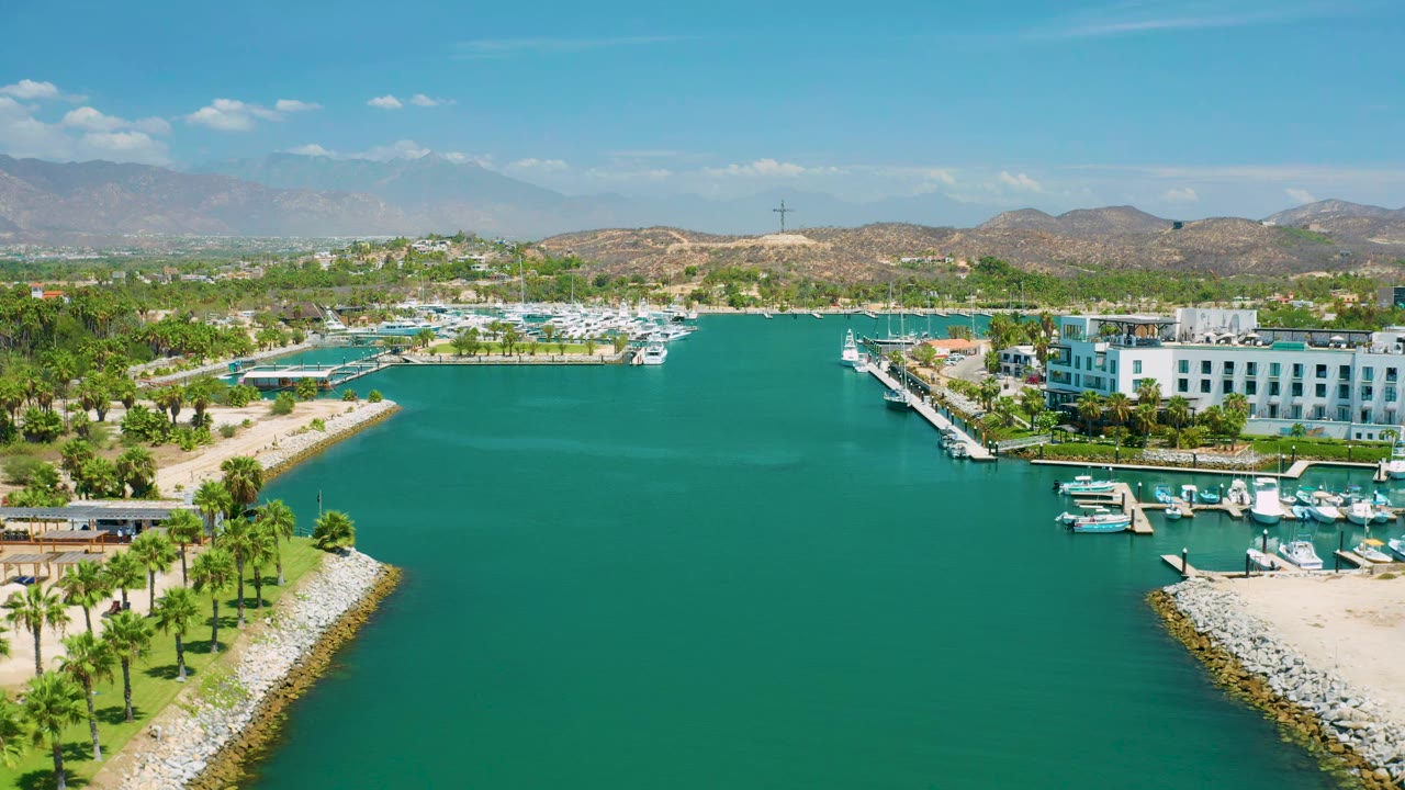 Second aerial frame from San Jose Del Cabo, Mexico with the green-blue water centered, a palm-lined jetty at lower right, marina docks and boats to the right, low buildings beyond, and no visible people.