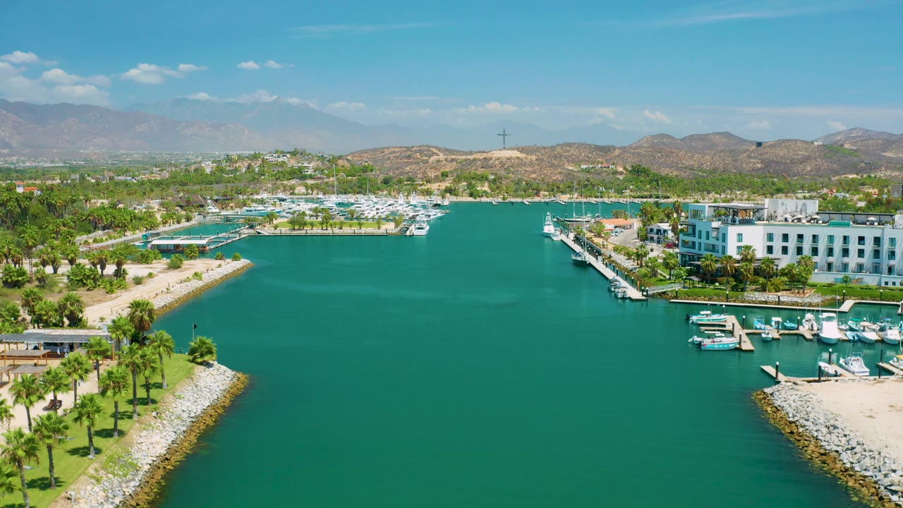 Aerial view in San Jose Del Cabo, Mexico showing the marina basin under bright light, with boats along the right-side docks, palms and rock edges below, hazy hills in the distance, and no visible people.