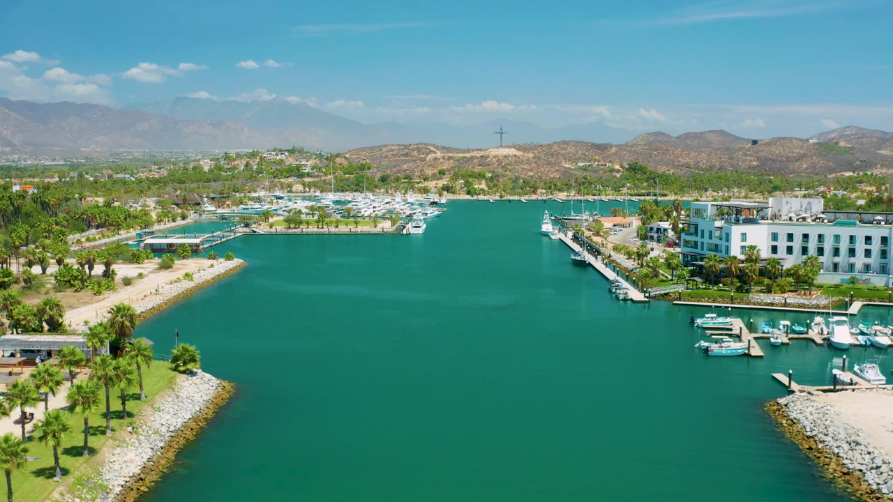 Mid-clip aerial frame from San Jose Del Cabo, Mexico with a broad water channel, palm-lined shore at left and lower right, marina docks and small boats near white buildings, distant brown hills, and no visible people.