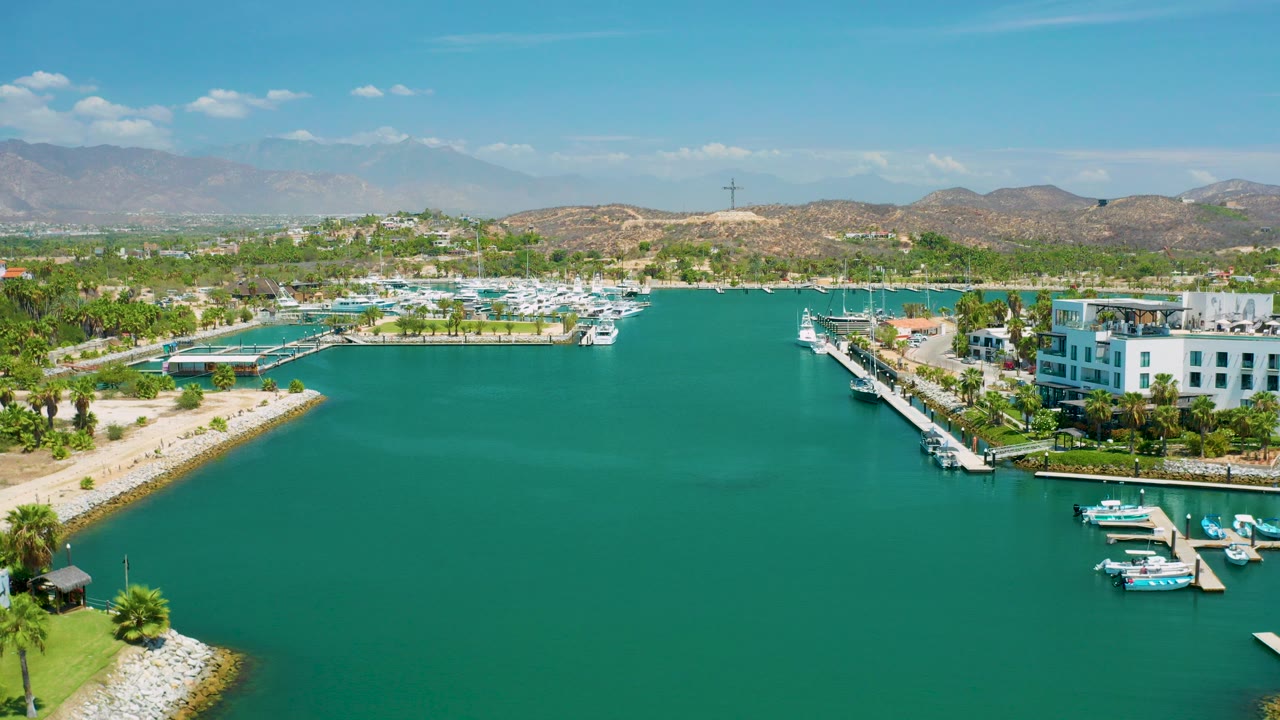 Opening aerial frame in San Jose Del Cabo, Mexico, looking across turquoise marina water toward docks, white resort buildings, palms, dry hills, distant mountains, and no visible people.