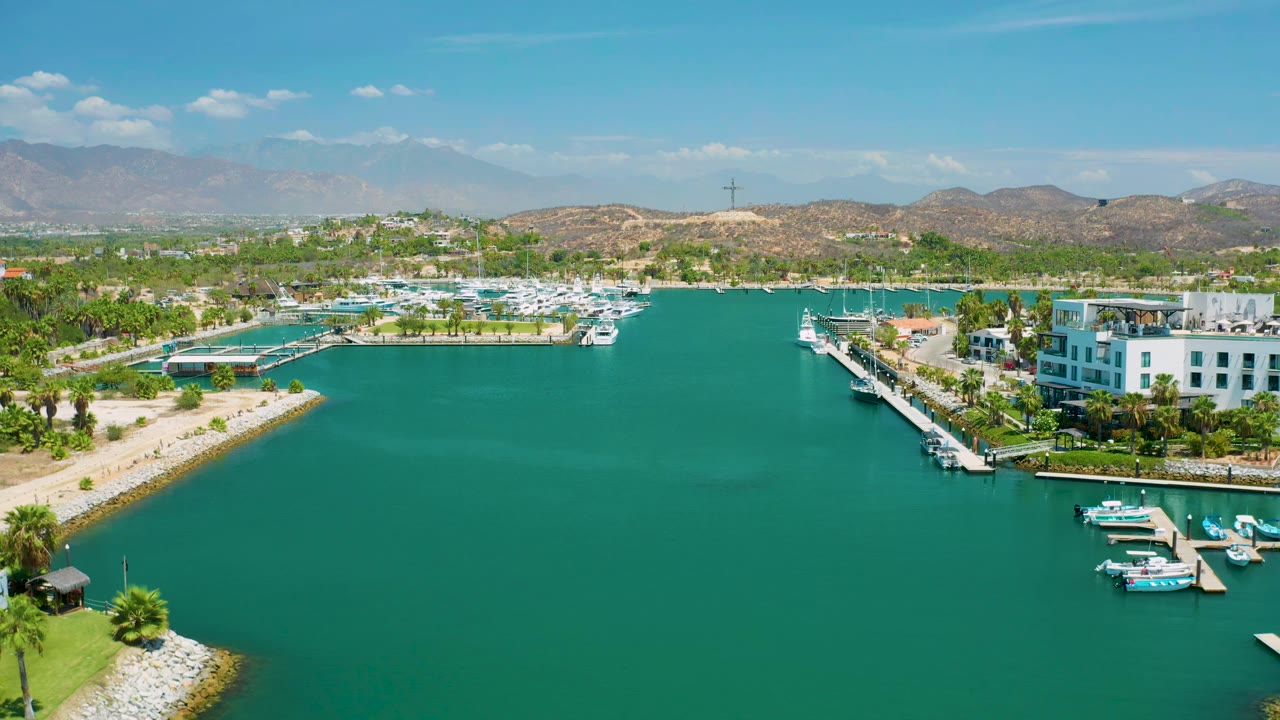 Second aerial frame in San Jose Del Cabo, Mexico, centered on the marina inlet with boats along docks, white waterfront buildings to the right, palms on both shores, mountains behind, and no visible people.
