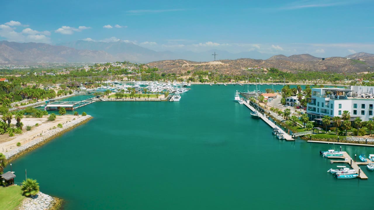 Third frame over San Jose Del Cabo, Mexico, with green-blue marina water dominating the foreground, a docked line of small boats at right, resort buildings at the edge, low hills and mountains in the distance, and no visible people.