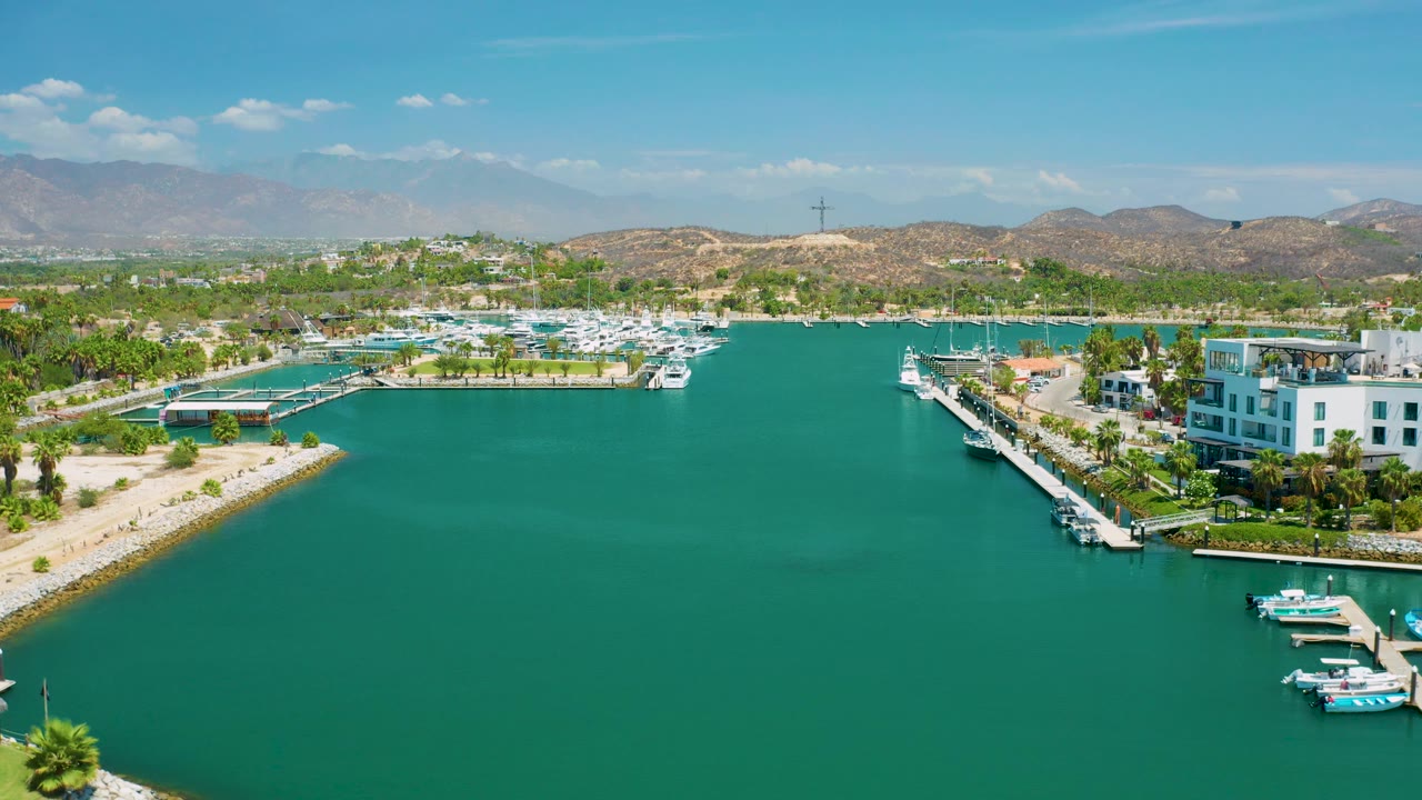 Resort marina viewed from above with open water, white buildings, docks, palms, hills, and mountains.