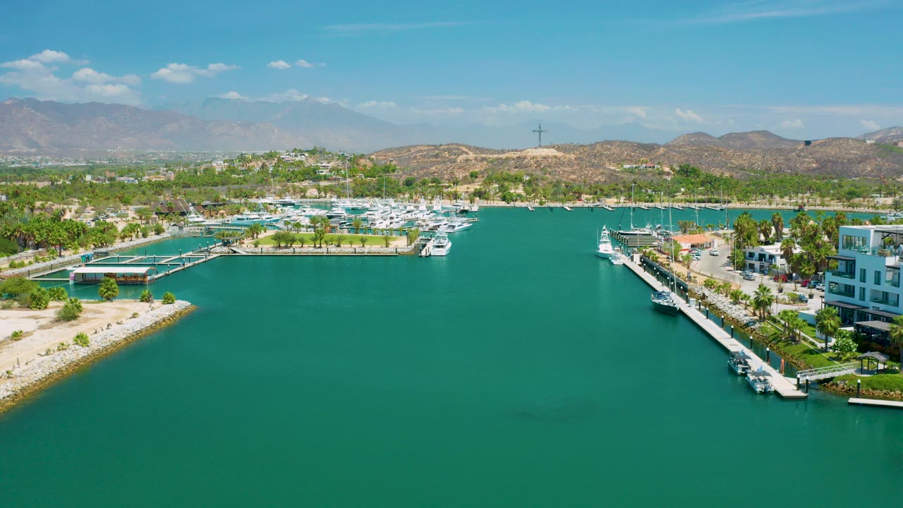 Aerial view in San Jose Del Cabo, Mexico, looking over turquoise marina water toward white boats, palm-lined docks, dry hills, distant mountains, and no visible people.