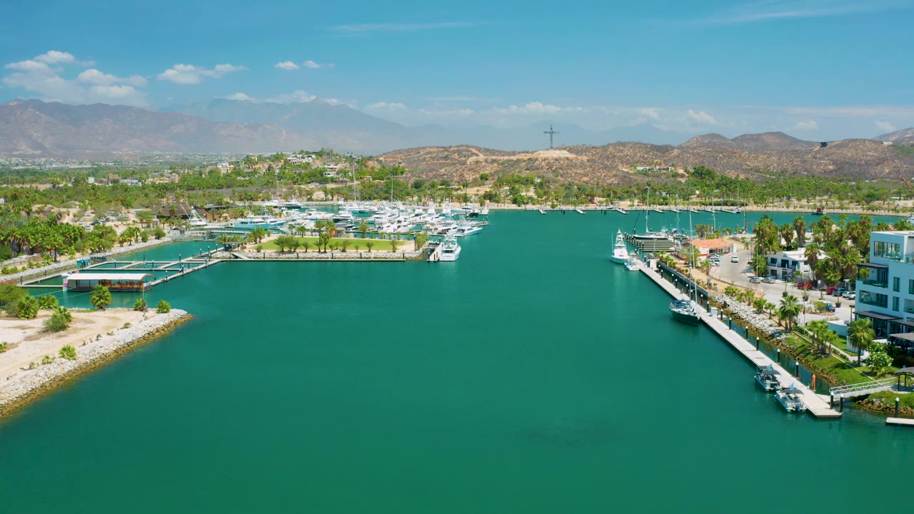 Wide aerial frame from San Jose Del Cabo places open turquoise water in front of marina docks, white boats, palm landscaping, arid hills, distant mountains, and no visible people.