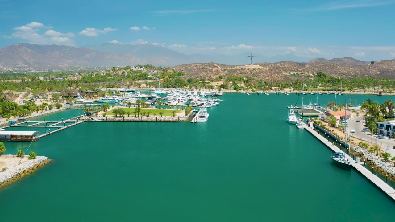 Drone-height frame of San Jose del Cabo marina water with a docked sailboat at the right edge, a small palm-lined peninsula below, distant hills, mountains, and no visible people.