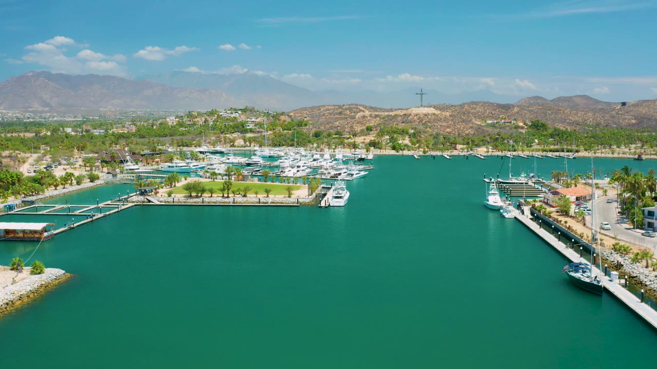 San Jose del Cabo aerial frame with broad open water in front, a marina and boats running across the midground, dry hills and faint mountains behind, and no visible people.