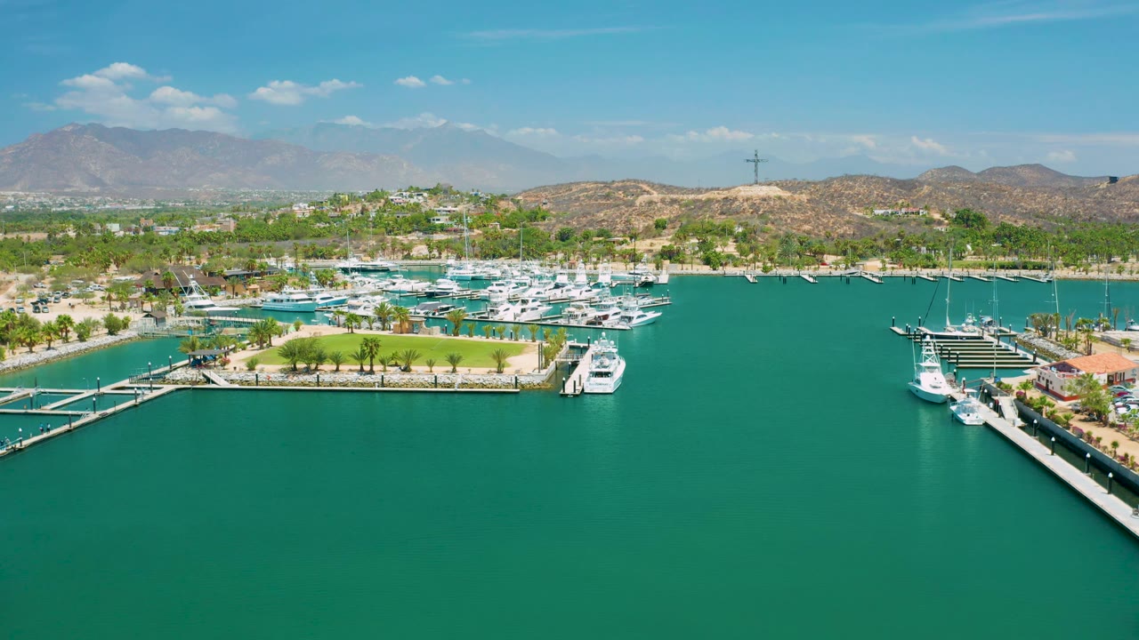 The marina basin in San Jose Del Cabo, Mexico, fills the frame with docked yachts, a small green point, palm trees, tan hills, distant mountains, light clouds, and no visible people.