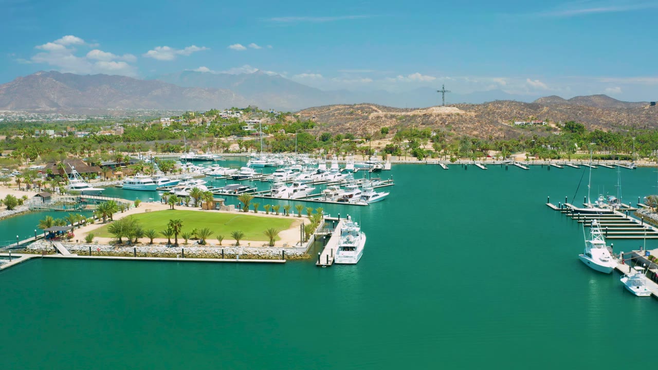 San Jose Del Cabo aerial frame with more of the curved green lawn in view, white yachts along docks, turquoise water, tan hills, mountains, blue sky, and no visible people.