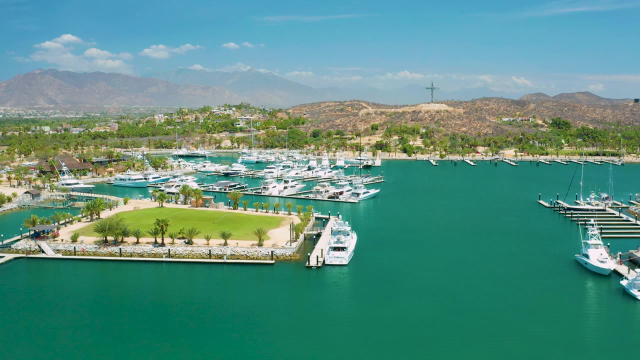 Aerial San Jose Del Cabo marina view with open turquoise water in the foreground, white boats clustered at docks, a lawn and palms at left, dry hills beyond, and no visible people.