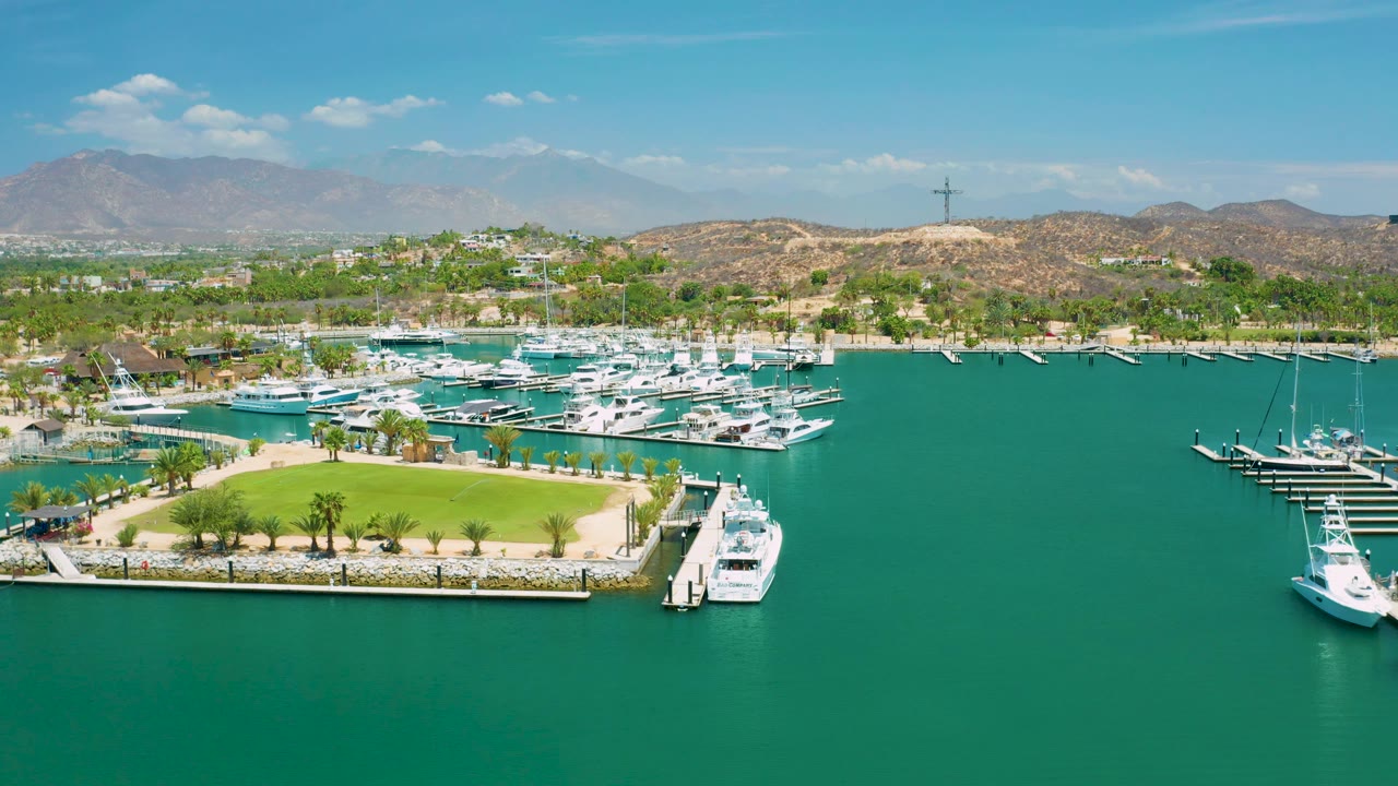 San Jose Del Cabo aerial frame with the green lawn low in frame, white boats packed into marina docks, turquoise water to the right, hills and mountains in the background, and no visible people.