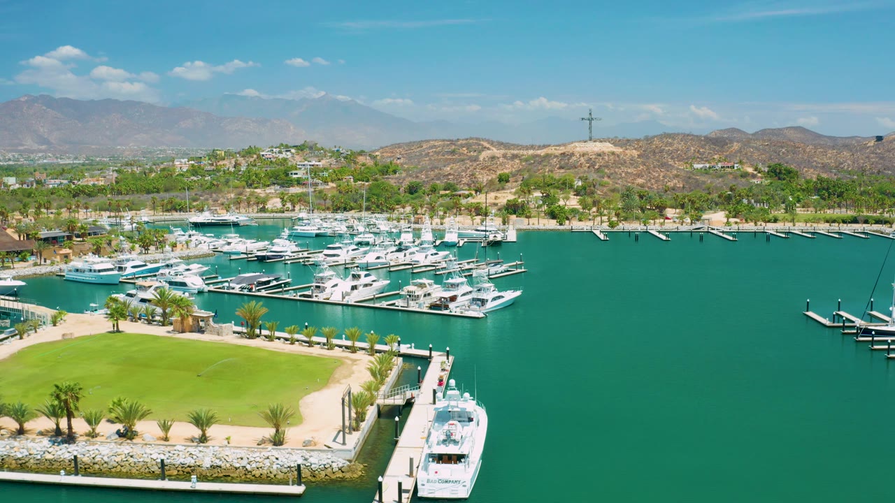 San Jose del Cabo marina frame with docked boats grouped on the left, a wide stretch of teal water on the right, the lawn and pier crossing the lower foreground, and no visible people.