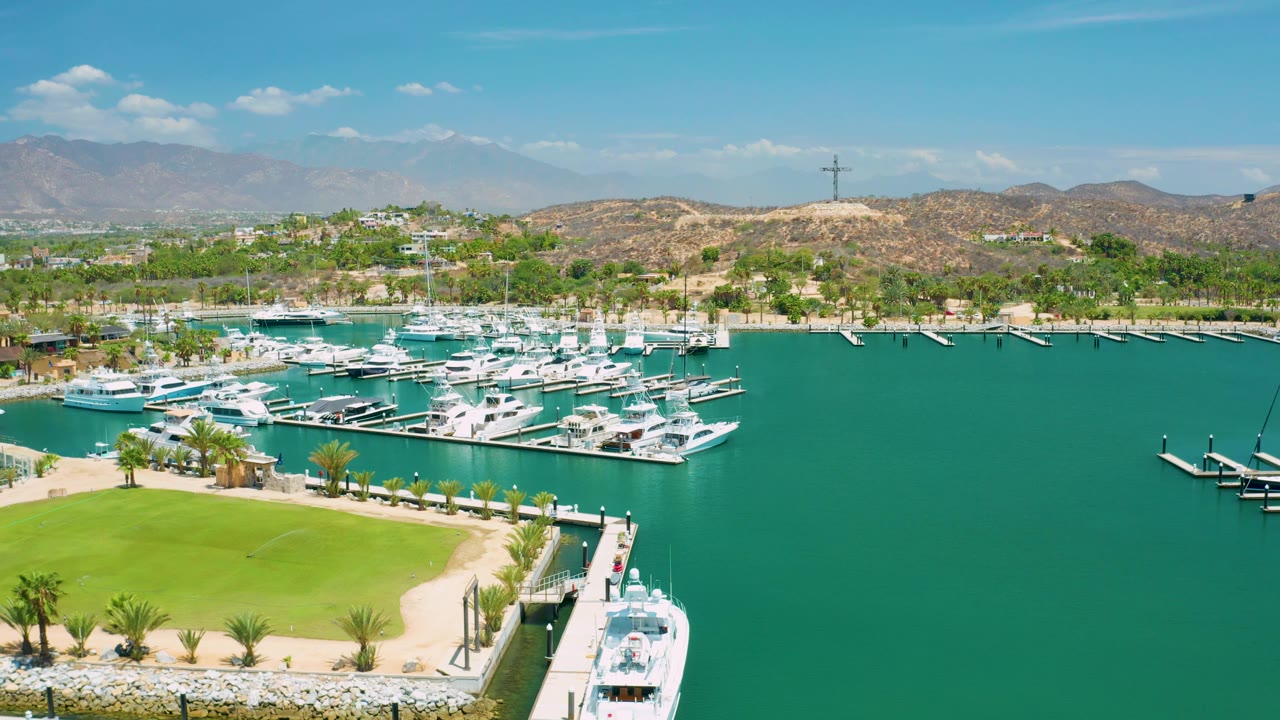 Wide aerial frame over the San Jose del Cabo marina with the green lawn filling the lower left, a white vessel near the lower right dock, clustered boats across the basin, and no visible people.