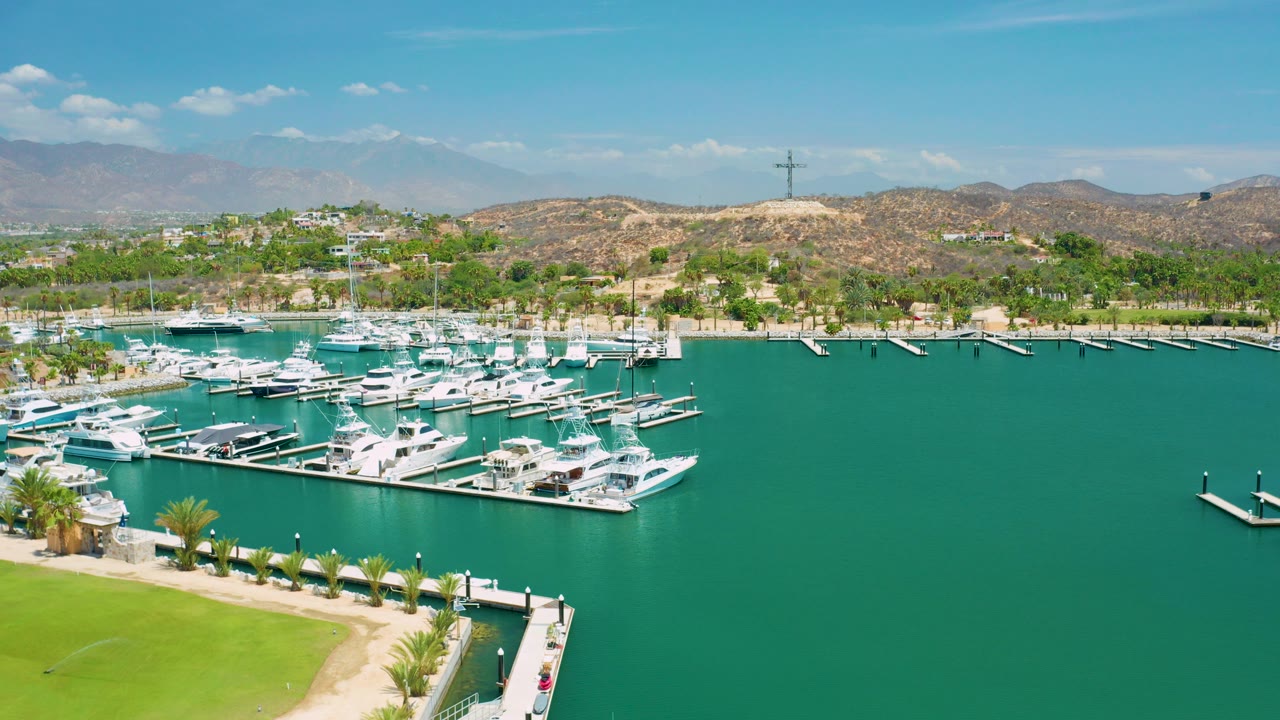 Opening aerial frame over a San Jose Del Cabo marina, with white yachts at floating docks, green water, a lawn edge, dry hills, blue sky, and no visible people.