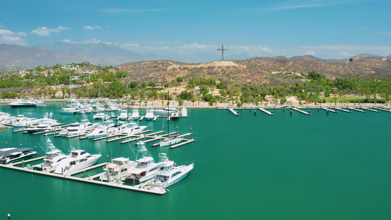 Aerial frame from San Jose Del Cabo showing white yachts clustered at foreground docks, green-blue marina water, a sandy shoreline, dry hills, distant mountains, and no visible people.