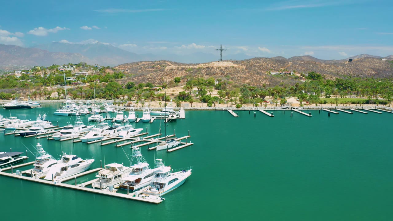 San Jose Del Cabo aerial frame showing yacht slips across the lower left, broad green-blue water across the center, a palm-lined shore, dry hills, distant mountains, and no visible people.