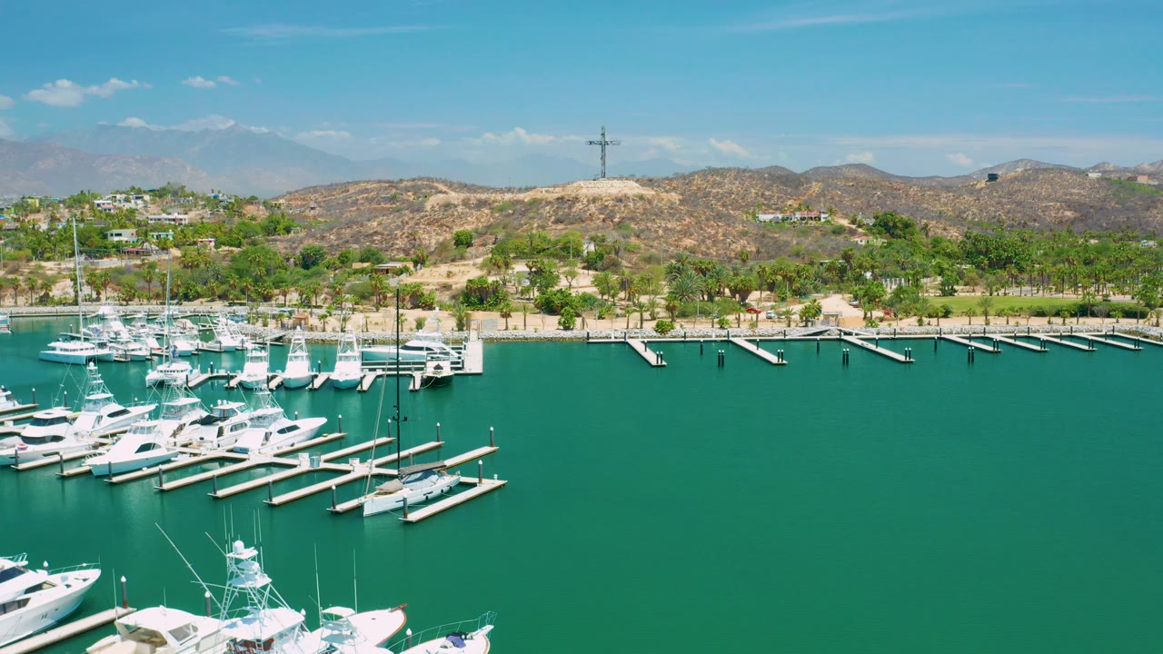 Aerial frame over San Jose Del Cabo marina showing white boats and docks in turquoise water, sandy shoreline, dry hills, distant mountains, blue sky, and no visible people.