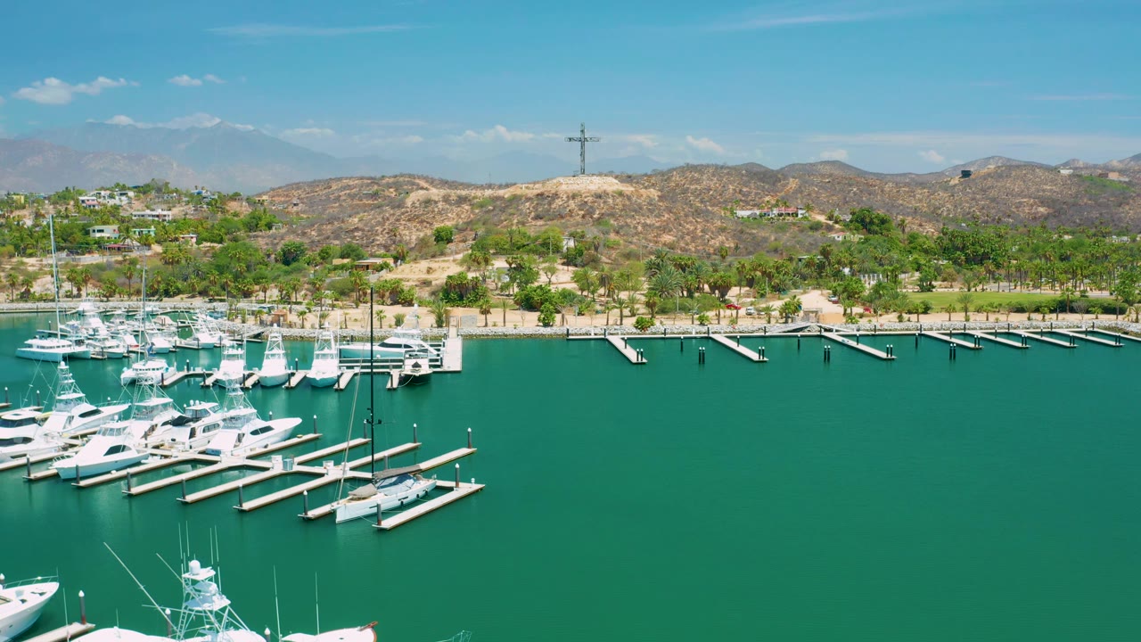 Wide San Jose Del Cabo marina frame showing open turquoise water in front, dock slips and white boats near the shore, palms and scrubby hills behind, and no visible people.