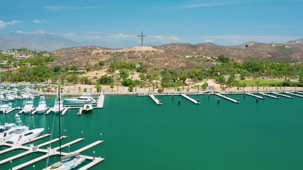 San Jose Del Cabo marina frame with several white yachts near the lower left, empty dock slips extending into green water, a palm-lined shore, tan hills, and no visible people.