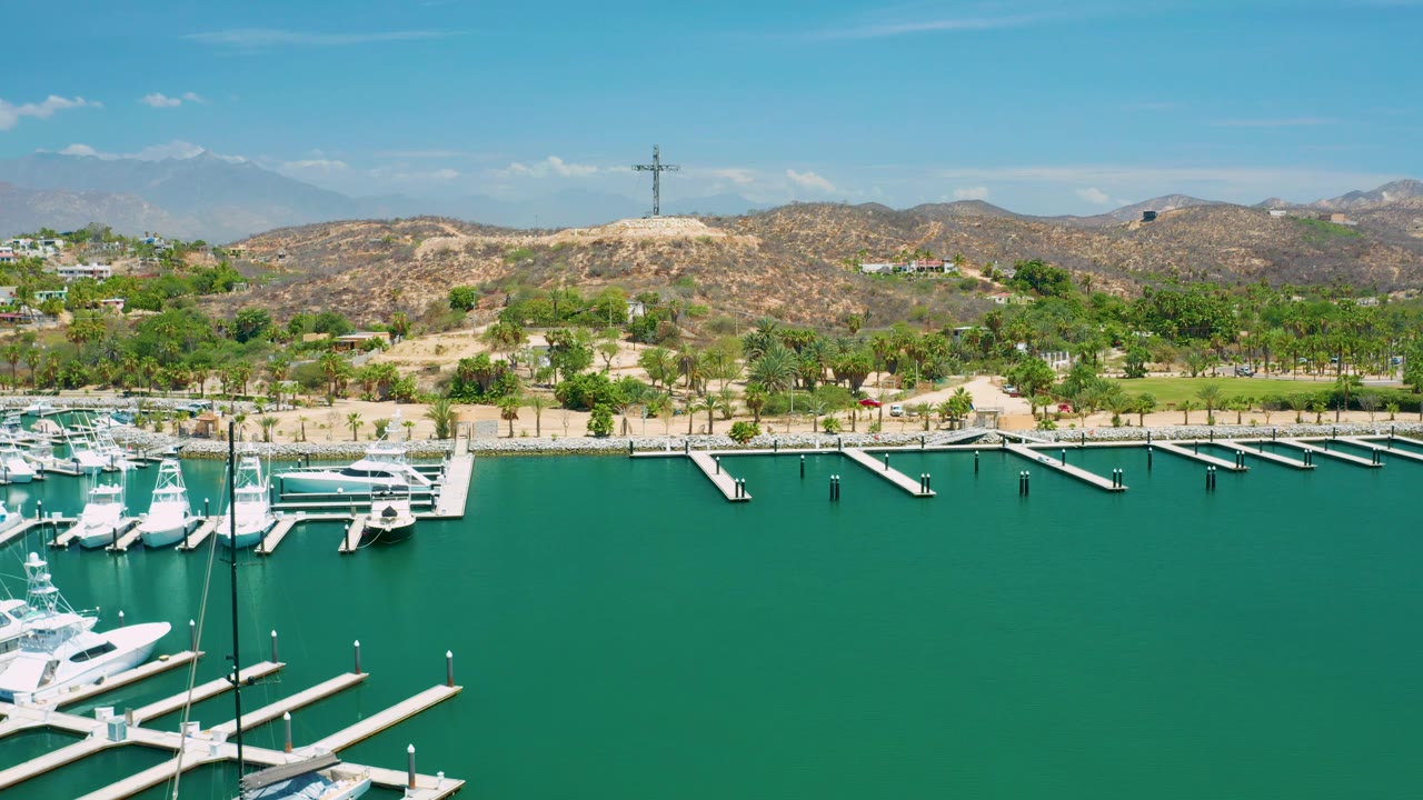 Aerial San Jose Del Cabo marina view with clustered white boats left of center, long dock fingers reaching into green water, palms along the shore, hills in back, and no visible people.