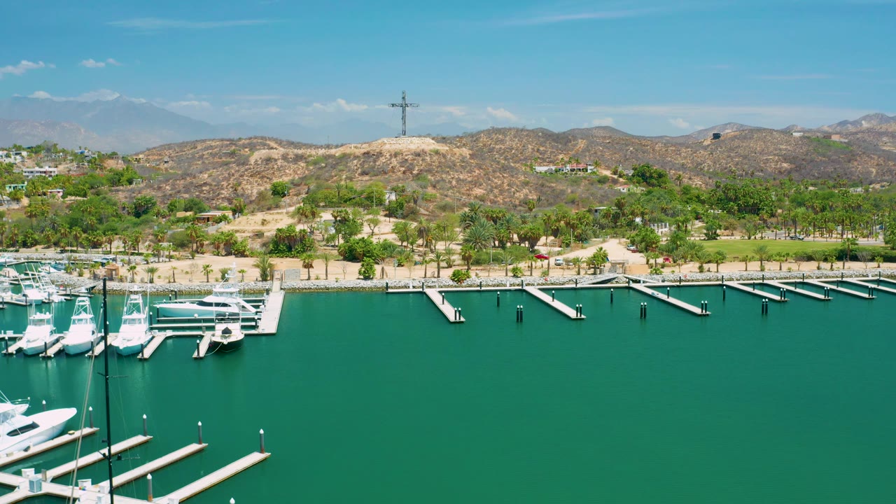 Wide San Jose Del Cabo marina composition showing open green water in the foreground, boats along the left edge, long docks across the midground, palm trees, dry hills, and no visible people.